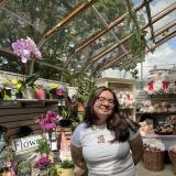 Jenn has long brown hair and glasses. She is wearing a white shirt with a small Teddy Bear on the  chest. She is standing in a green house with flowers and teddy bears on the shelves. The sun is shining through the greenhouse ceiling, there is a tree outside and the sky is bright blue with puffy white clouds.